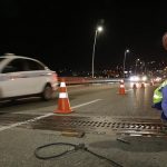 dois trabalhadores usando vestimentas de proteção aparafusam placa de metal no chão da ponte com cones em volta, à noite, e taxi passando ao lado