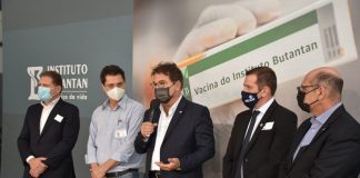 em assinatura de intenção de compra, cinco homens em pé lado a lado; painel com vacina ao fundo; um deles fala ao microfone; todos de máscara
