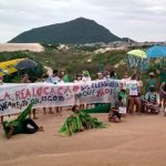 Moradores do Santinho protestam com faixa "pela realocação das elevatórias de esgoto"; alguns vestidos de jacaré; morro das aranhas e dunas ao fundo