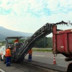 maquina recortando asfalto da via expressa sul e jogando detrito em caçamba de caminhão; operários ao lado; trânsito ao fundo