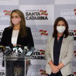 daniela reinehr e carmen zanotto usando máscara, em pé, em frente a bancada com microfones; ao fundo painel com logos do governo de santa catarina