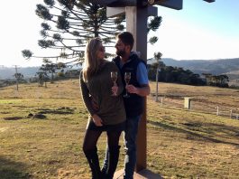 Roberta Zilli e Bruno Buske olham um para o outro em paisagem serrana segurando taça de vinho